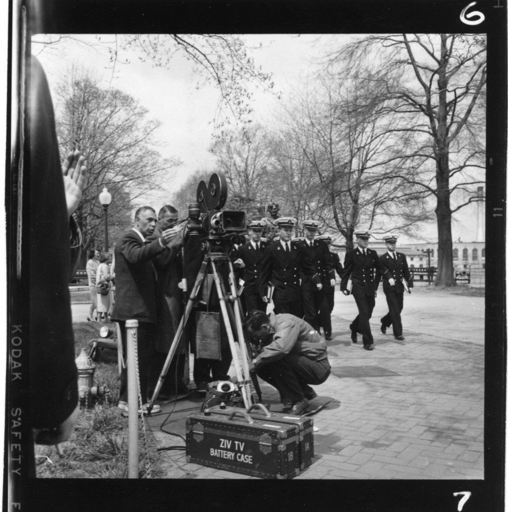 A production still shows a 1950s-era film camera with a case labeled "Ziv TV battery case" on the ground in front of it. A group of actors dressed as sailors are positioned behind the camera, about to walk into frame.