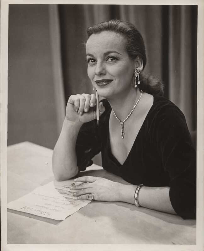 A white woman sits at a table and gazes into the distance slightly above and beyond the camera with a soft, pleasant expression as though she is recalling a fond memory. She wears a black dress with a deep v-neck, and her light hair is elegantly pulled back. Her sparkling necklace and elaborate earrings appear to drip off her person like icicles, and she holds a pencil in her right hand while her left hand touches a paper list in front of her on the table. 