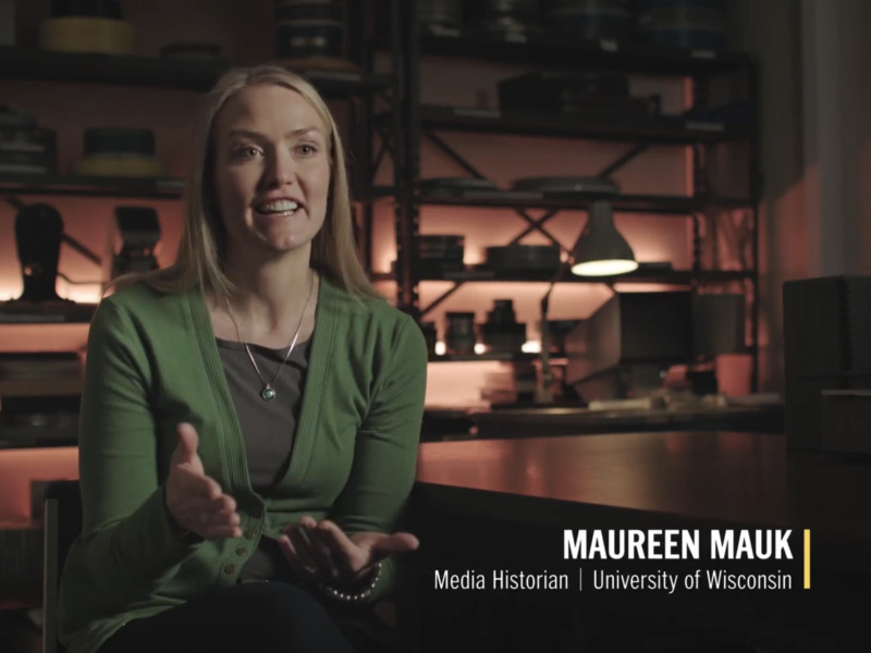 A blonde-haired white woman wears a green cardigan over a gray shirt. She is sitting in a darkened room in front of various film preservation and archive tools. A chyron names her as "Maureen Mauk, media historian, University of Wisconsin."