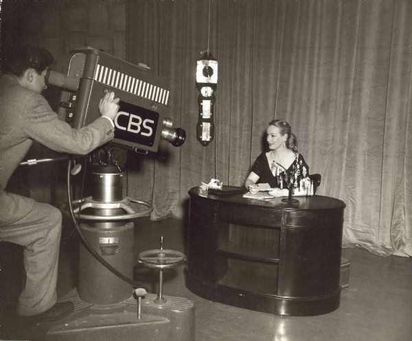 A blonde woman sits at a wooden desk. A video camera labeled CBS is pointed at her.