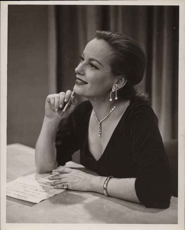 A white woman sits at a table and gazes into the distance to the far left of the camera with a soft, pleasant expression as though she is recalling a fond memory. She wears a black dress with a deep v-neck, and her light hair is elegantly pulled back. Her sparkling necklace and elaborate earrings appear to drip off her person like icicles, and she holds a pencil in her right hand while her left hand touches a paper list in front of her on the table. 