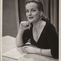 A white woman sits at a table and gazes into the distance slightly above and beyond the camera with a soft, pleasant expression as though she is recalling a fond memory. She wears a black dress with a deep v-neck, and her light hair is elegantly pulled back. Her sparkling necklace and elaborate earrings appear to drip off her person like icicles, and she holds a pencil in her right hand while her left hand touches a paper list in front of her on the table. 