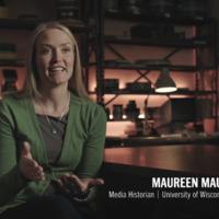 A blonde-haired white woman wears a green cardigan over a gray shirt. She is sitting in a darkened room in front of various film preservation and archive tools. A chyron names her as "Maureen Mauk, media historian, University of Wisconsin."