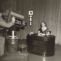 A blonde woman sits at a wooden desk. A video camera labeled CBS is pointed at her.