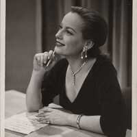 A white woman sits at a table and gazes into the distance to the far left of the camera with a soft, pleasant expression as though she is recalling a fond memory. She wears a black dress with a deep v-neck, and her light hair is elegantly pulled back. Her sparkling necklace and elaborate earrings appear to drip off her person like icicles, and she holds a pencil in her right hand while her left hand touches a paper list in front of her on the table. 